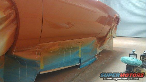 Orange car body panel covered in masking tape and plastic sheeting, with blue and yellow tape visible