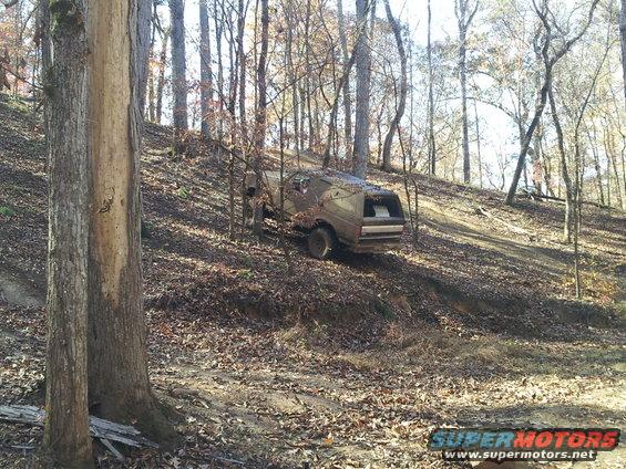 Brown SUV with a cap driving up a dirt hill in a forest. Appears to be a 1980s model