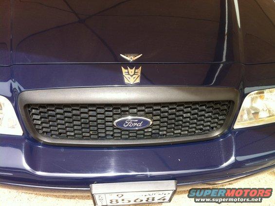 Dark blue Ford Crown Victoria front end with a honeycomb grille, Ford logo, and Transformers Autobot and Decepticon symbols on the hood