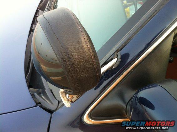 Close-up of a car's side mirror with a dark leather cover and stitching. A chrome trim piece is visible along the door frame