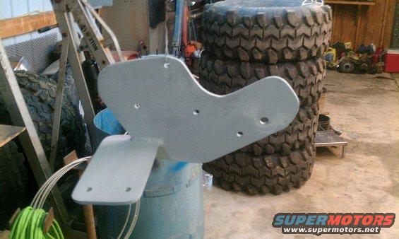 Unpainted metal bracket with three mounting holes, resting on a blue bucket. Stack of tires in background