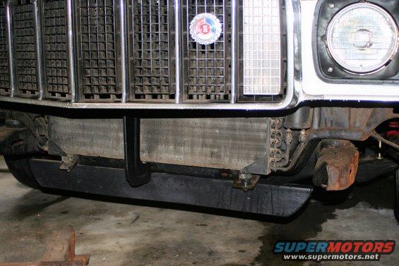 Front of a vehicle showing grille, radiator, and part of a headlight. A circular badge is on the grille