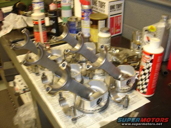 Engine pistons and connecting rods laid out on newspaper with various automotive fluids and cleaning supplies in the background