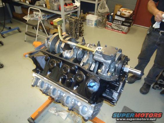 Engine block on an orange stand, showing crankshaft, pistons, and connecting rods. A person stands in the background