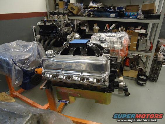 V8 engine on an orange stand, with another engine covered in plastic behind it. Shelves with parts are in the background