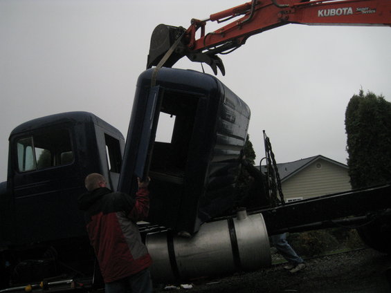 Dark blue truck cab being lifted by an orange excavator arm, with a man in a red jacket assisting