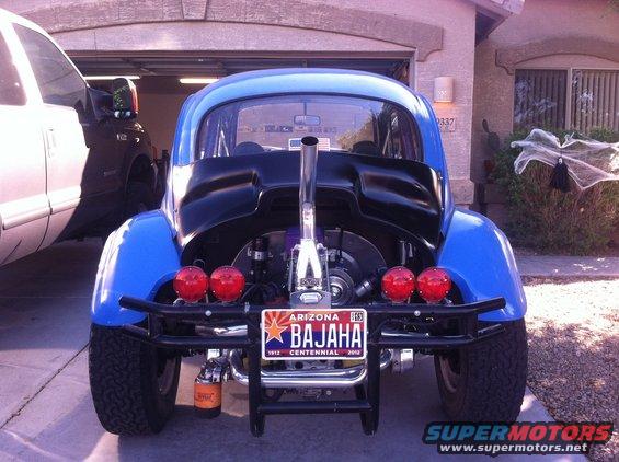 Rear view of a blue custom VW Beetle with a large exhaust pipe and off-road tires. A white pickup truck is parked next to it