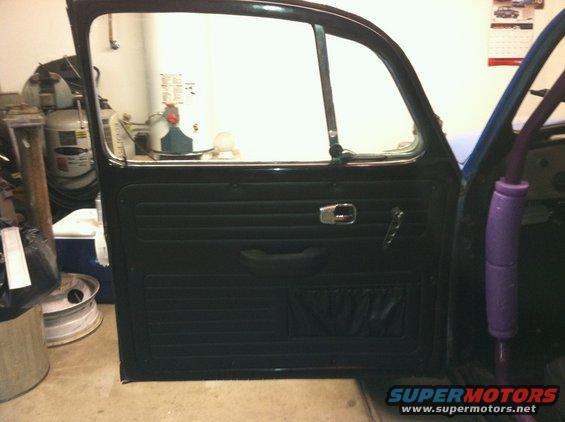Interior door panel of a classic VW Beetle, showing black upholstery, chrome handle, and window crank