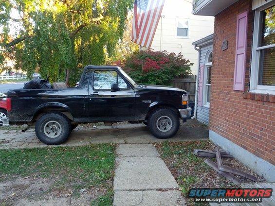 Black 1995 Ford Bronco parked next to a brick house with an American flag flying