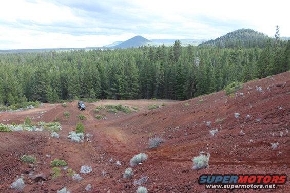 SUV on red dirt road with pine forest and mountains in background