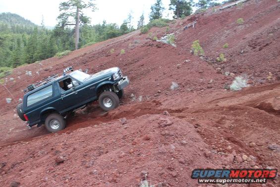 Dark green Ford Bronco climbing a steep, red dirt hill with trees in the background