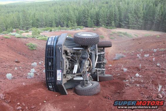 1995 Ford Bronco overturned on its side in a dirt landscape with trees in the background. License plate reads EB HJ