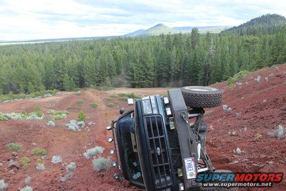 Dark green Ford Bronco flipped on its side on a red dirt hill, spare tire visible