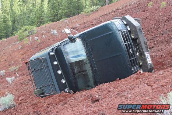 dark truck tipped on its side in red dirt with trees in background