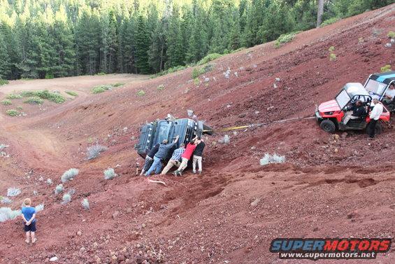 People pushing a stuck off-road vehicle on a red dirt hill while another side-by-side vehicle pulls it with a strap