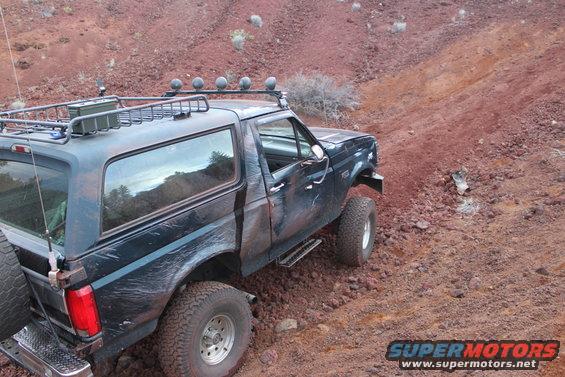 Dark blue Ford Bronco with roof rack and off-road tires driving on a red dirt hill
