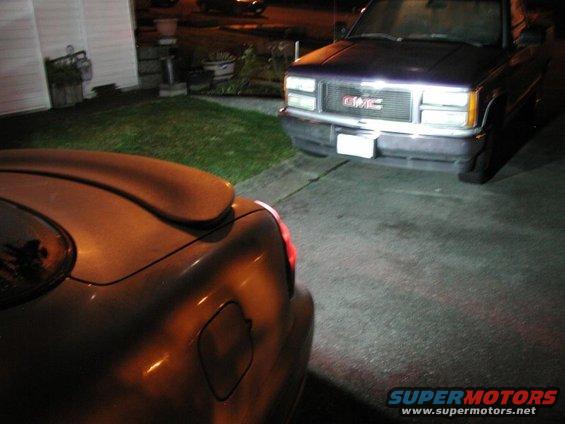 Rear of a car with a spoiler, parked in front of a GMC pickup truck at night