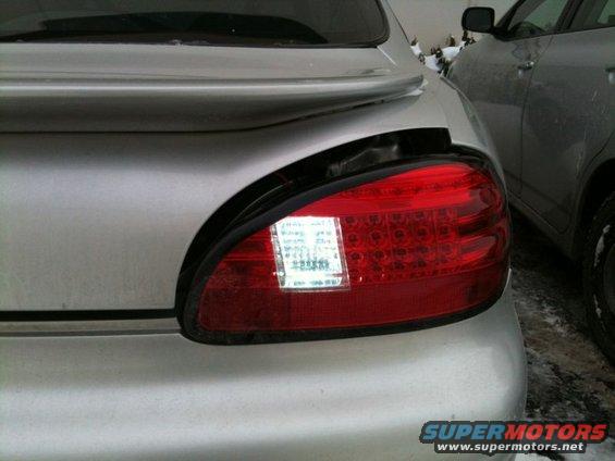 Silver sedan's damaged rear tail light assembly with visible LED elements and a white sticker. Another vehicle is parked to the right