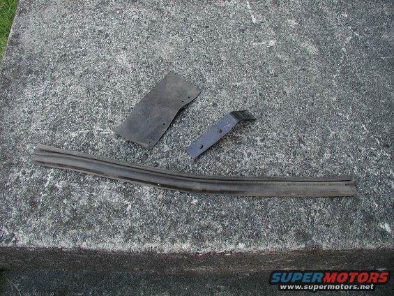 Three black car parts, including a rubber seal and two metal brackets, lie on a concrete surface