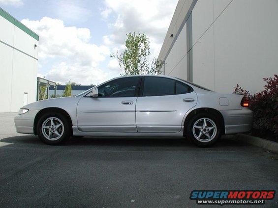 Silver sedan parked next to a building, side view. The car has tinted windows and five-spoke wheels