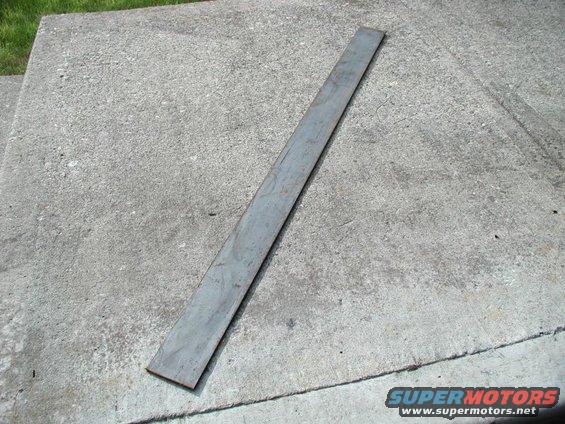 Long flat gray steel bar stock lying diagonally on a concrete driveway