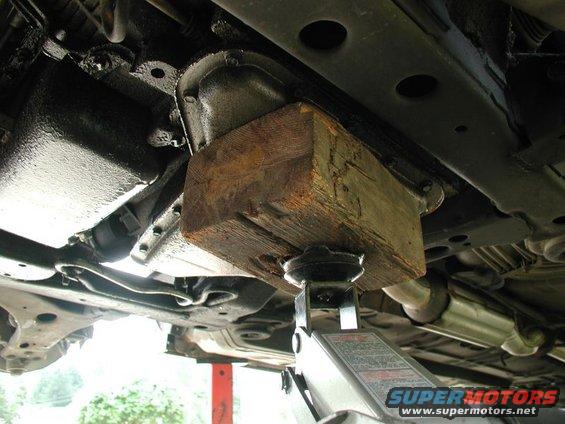 Underside of a vehicle supported by a jack and wooden block, showing the transmission and exhaust system