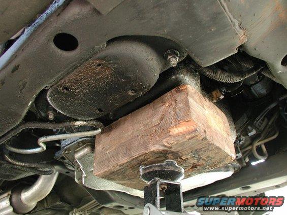 Underside of a car supported by a jack and a wood block. Shows frame, exhaust, and some wiring