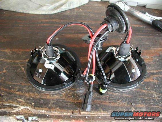 two black fog lights with red and black wires connected, sitting on a wooden surface