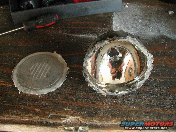 Disassembled headlight assembly with reflector and cover on a wooden surface. Tools are visible in the background