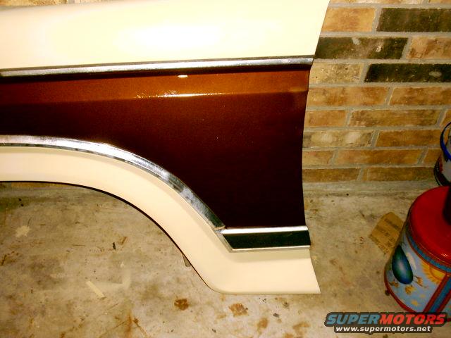 Close-up of a car fender with two-tone paint and chrome trim, against a brick wall