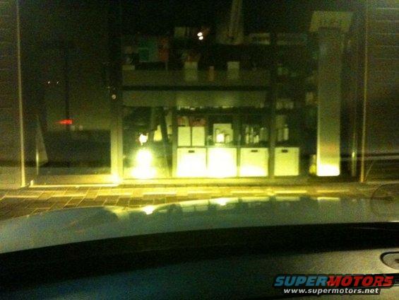 View from inside a car looking out at a brightly lit store at night. The store has shelves stocked with items