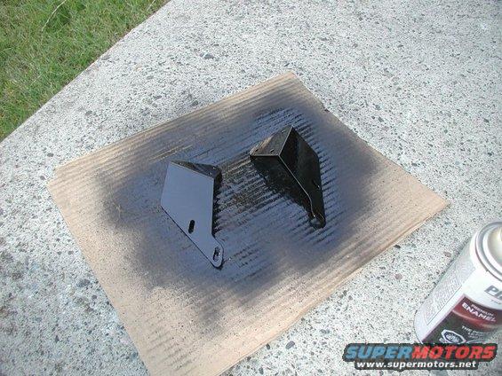 Two black metal brackets on cardboard with spray paint overspray and a can of spray paint