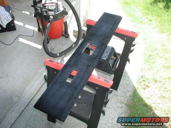 Black carpeted overhead console part with cutouts, resting on sawhorses next to a red shop vac
