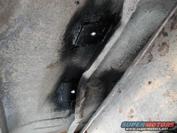 Close-up of a vehicle's underbody showing a square metal plate with two bolts, possibly for seat mounting