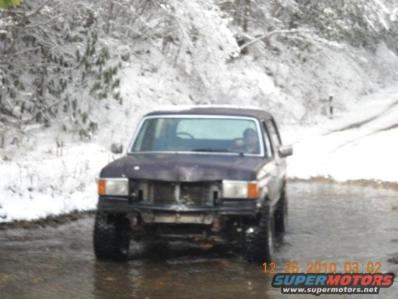 Dark purple Ford Bronco with missing grille and front bumper, driving through water on a snowy road