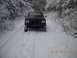 1990 Ford Bronco - A black SUV drives down a snow-covered road surrounded by trees.