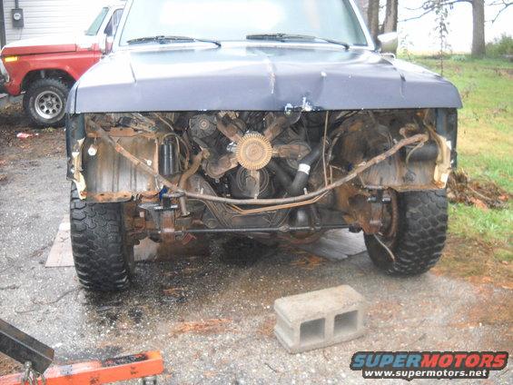 Front view of a 1990 Ford Bronco with grille removed, showing engine. Another red truck is visible