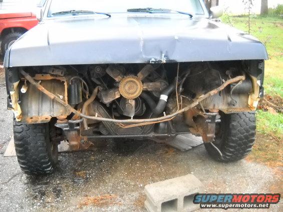 Truck front end missing grille and bumper, showing engine and radiator. Red truck in background