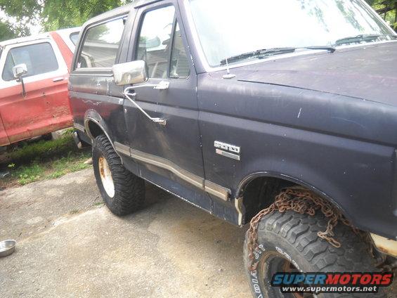 Dark blue Ford Bronco with a dented driver's side door and chains on the front tire