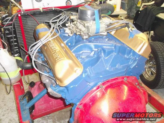 Blue Ford V8 engine with gold valve covers on a red stand, carburetor visible
