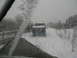 1987 Ford Bronco - Two trucks drive down a snow-covered road, viewed from inside another vehicle during a snowstorm.