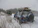 1987 Ford Bronco - Two vehicles, a blue SUV and a military truck, navigate a snowy landscape with trees in the background.