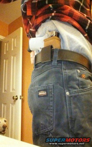 Person's hip area showing a concealed handgun in a holster, dark wash jeans with pockets, and a plaid shirt