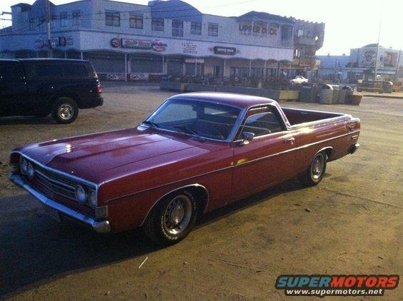 Red 1968 Ford Ranchero parked on asphalt, with a black SUV to its left and buildings in the background