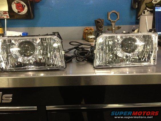 Two aftermarket projector headlights sit on a metal workbench. Wires are attached to the left headlight