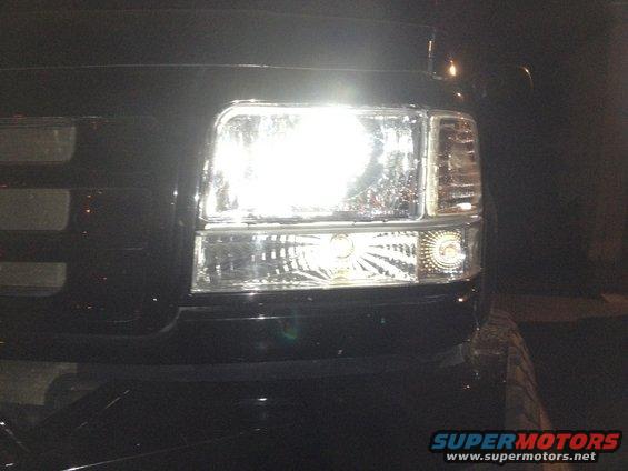 Close-up of a vehicle's headlight assembly with the light on. Watermark reads SUPERMOTORS