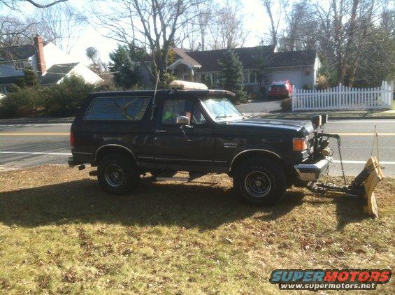 Black Ford Bronco with snow plow attached, parked on grass next to a road