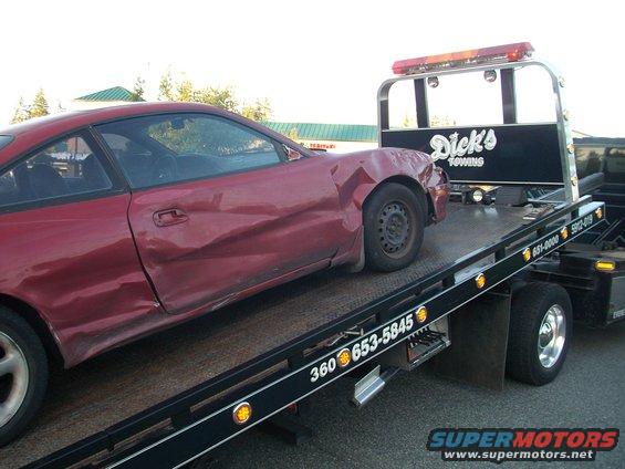 Side view of a damaged red 1989 Ford Probe on a tow truck. The car has significant damage to the front fender and driver's side door
