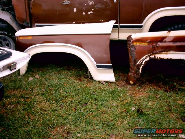 rusted brown vehicle fender with white trim, sitting on grass next to another vehicle part