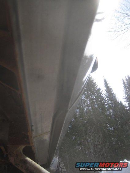 Truck body panel with visible rust and a curved edge, seen from a low angle against a backdrop of trees
